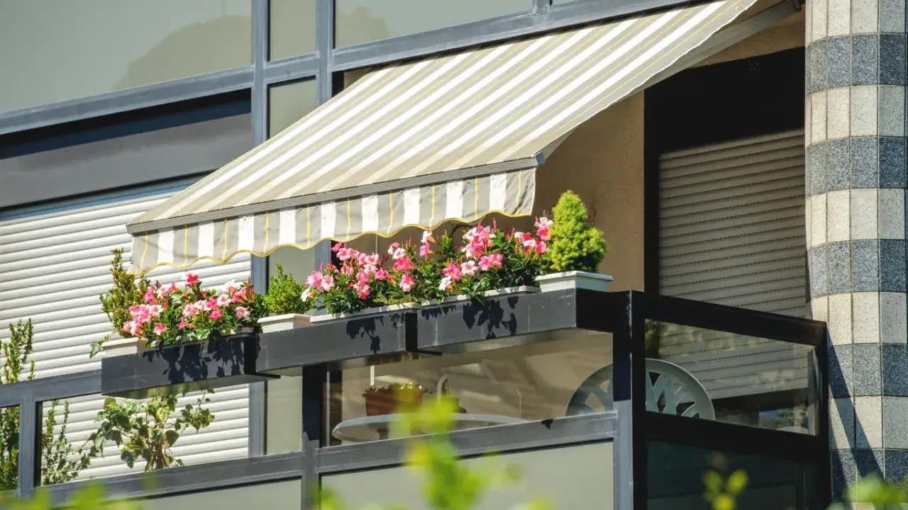 Markiza nad balkonem w bloku, z kwitnącymi różowymi kwiatami i zieloną rośliną, to świetny sposób, jak osłonić balkon.